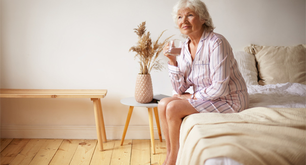 elderly lady sitting on the edge of the bed with feet on the floor holding a glass of water